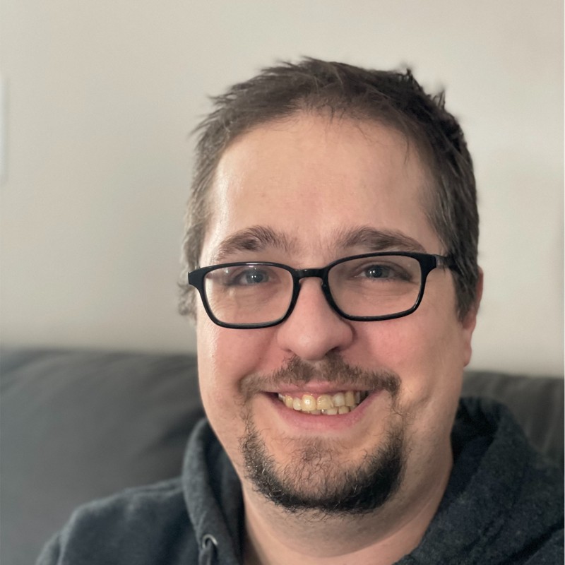 Ryan Sullivan smiling, he has brown hair, hazel eyes, a goatee, and wears glasses.